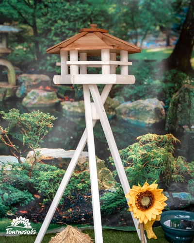 Mangeoire sur pied en bois blanc au toit rouge-brun, avec un tournesol décoratif, devant un décor de jardin japonais
